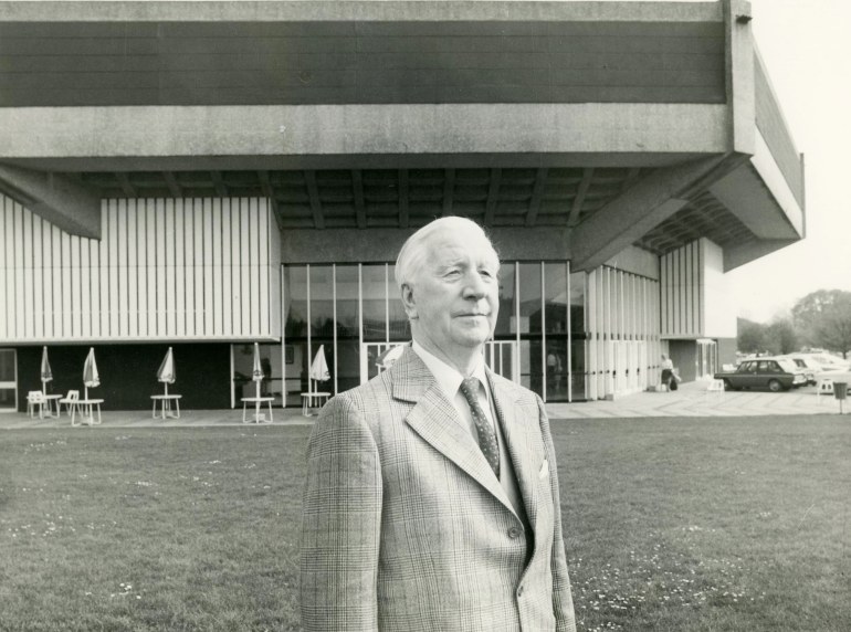 A sepia portrait of Leslie Evershed-Martin standing on the green outside the Main House of the Chichester Festival Theatre.