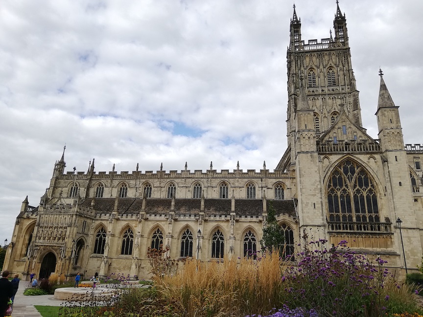 Exterior photograph of Gloucester Cathedral, showing the main entrance, bell tower and gardens