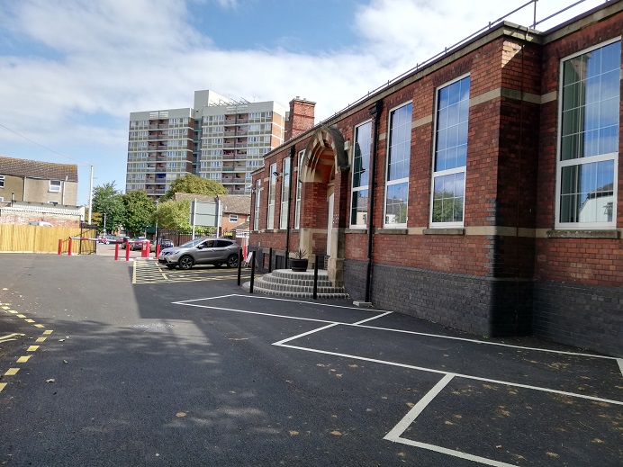 Showing the side of the building with assorted car parking spaces. The building is red and grey brick, with large rectangular windows.