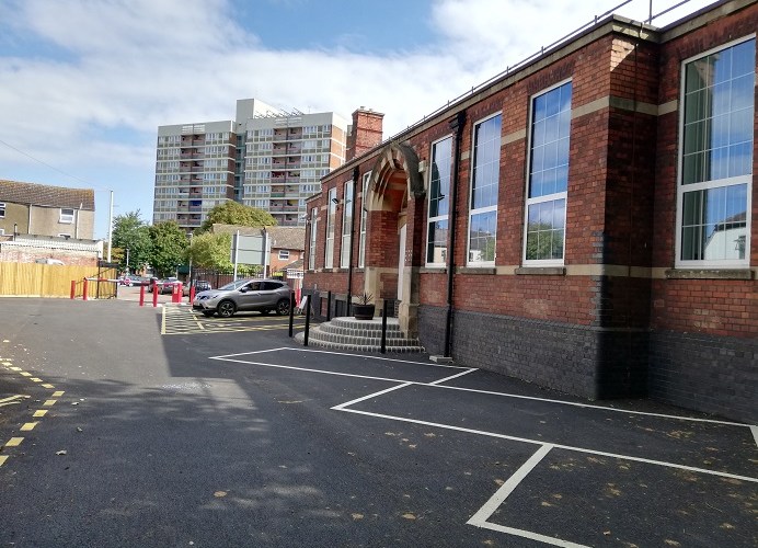 Showing the side of the building with assorted car parking spaces. The building is red and grey brick, with large rectangular windows.