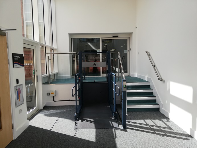 Stairs and a wheelchair lift bring you up to automatic door. The carpets are light grey and teal green. A side door leads to a community centre, and another side door leads to the garden