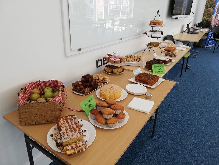 Apples and cakes line two tables. Including brownies, lemon drizzle, victoria sponge, cupcakes, marmalade cake, coffee and walnut, chocolate cake, chocolate and vanilla and fruit cake.