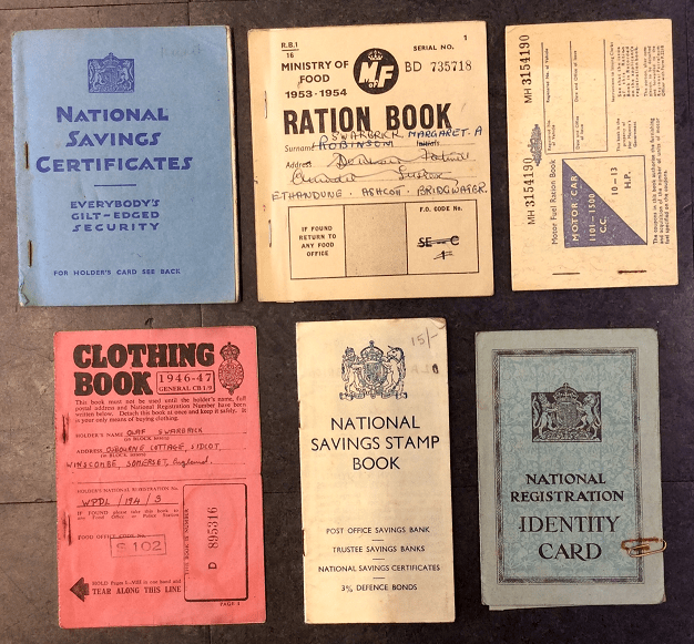 Six booklets held by a member of the public during the rationing period, including a blue National Savings Certificates booklet, a Ration book, a Petrol Ration book, a red Clothing Ration book, a slim National Savings Stamp Book, and a blue National Registration Identity Card