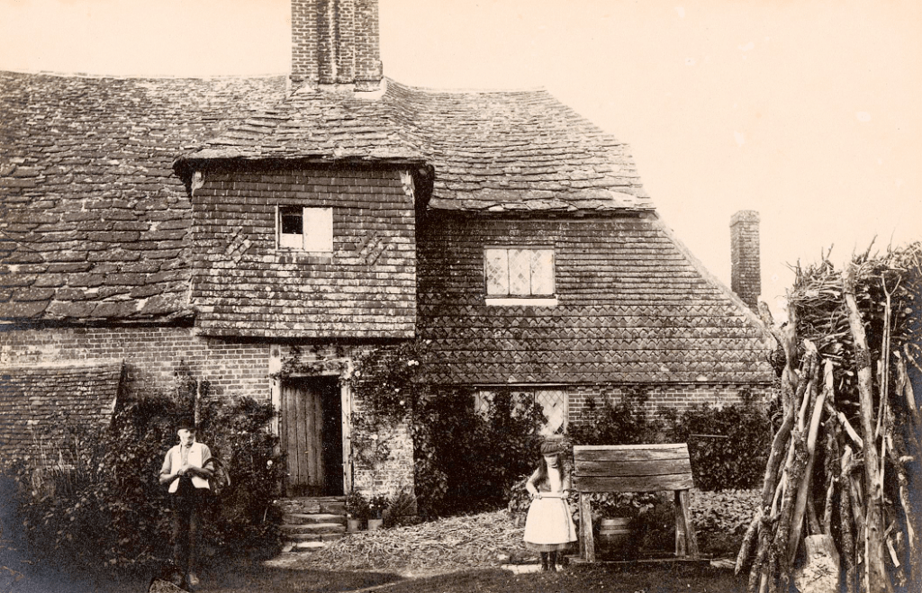 A sepia image of a tiled cottage. There is a little girl standing next to well and a man an standing next to doorway.