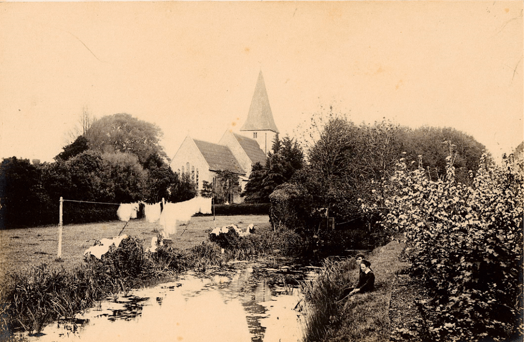Sepia image of a river, with church in background. Two young boys sitting on bank, washing hung on line by river