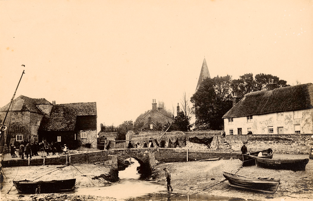 Sepia image of a brick bridge surrounded by buildings, boats and people. There is a church visible in background. There are also nets drying on stone walls