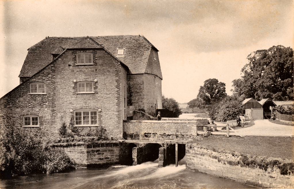 Sepia image of a side view of mill and brick bridge over the river. Man walking over bridge and one man kneeling against a wooden fence.