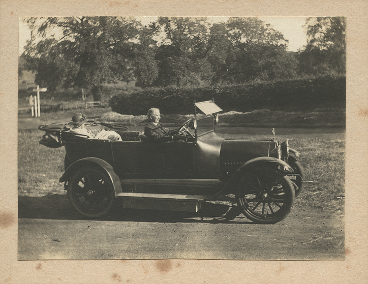 Side view of soft top car with the roof down, Carruthers at the wheel with Beatrice in the rear
