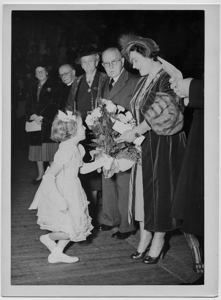 Corfield standing next to the Queen Mother who is being presented with a bouquet of flowers by a curtsying young girl