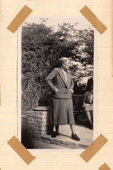 A photograph of Nancy Price taken from her scrapbook of work for the South Downs. Shows Nancy posing with her hands in her jacket pockets, standing in front of her wooden bench in High Salvington.