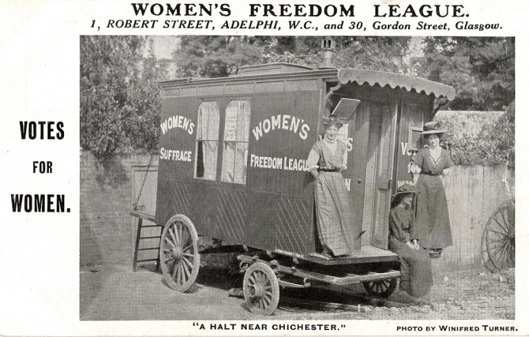 Two women are standing on rear step of the caravan with a third, seated woman (unidentified). Women's Suffrage and Women's Freedom League are written on the side of the caravan.
