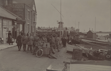 A dozen or so men in uniform and women and children posing for the photograph. Some are sitting on the floor, some are smilling.
