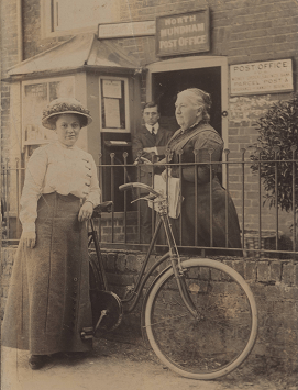 Three members of staff, two women and one man, standing in Edwardian clothes in front of the post office. A bike rests against a fenced wall and one woman.