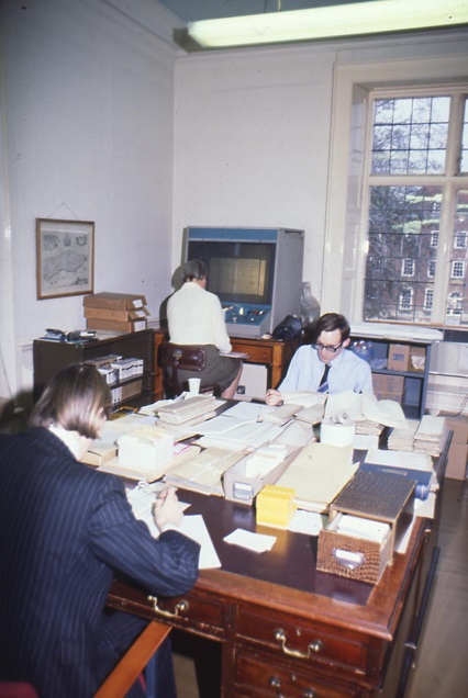 Both working at large desks surrounded by index card drawers and papers, another member of staff is working at a large microfilm reader in the background