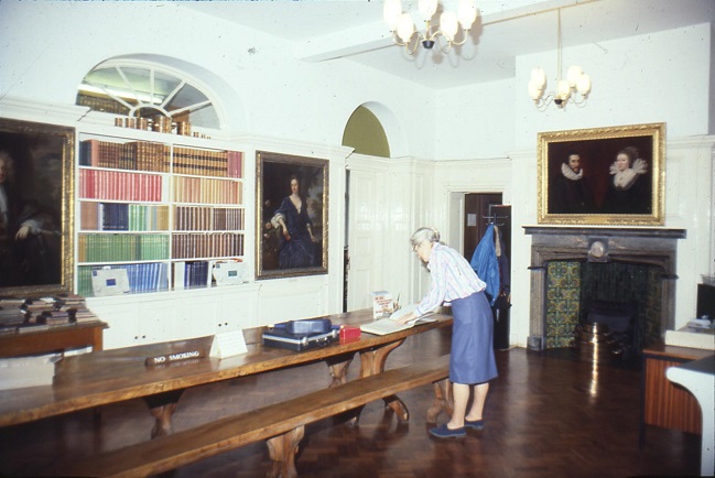 Portraits hanging on walls and books on shelves, visitors book is kept on a large, refectory-style, highly polished wooden table, tiled fireplace visible in background