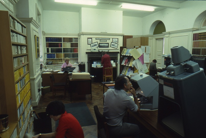 Researchers using microfilm readers, microfilm storage unit behind them, Receptionist at desk in far corner
