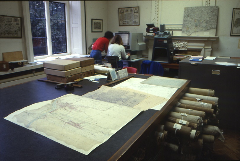 Maps on the large desk, rolled maps protruding from storage built into the desk, two people using a microfilm reader in the background