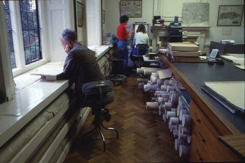 Researcher working on large windowsill with rolled maps stored in a recess beneath him, rolled maps stored in built-in storage in the map table, two women using a microfilm reader in the background)