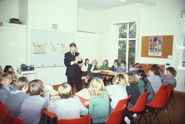 School children sitting on chairs in a horseshoe shape around Kim Leslie, who is holding up a document