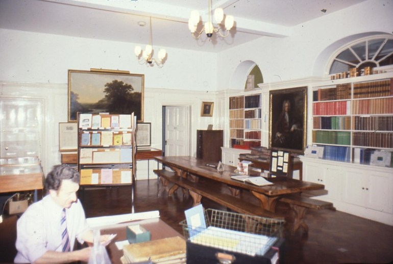 Shows large, highly polished refectory-style table, leaflets on stand, paintings and portraits on wall and books on built-in shelves