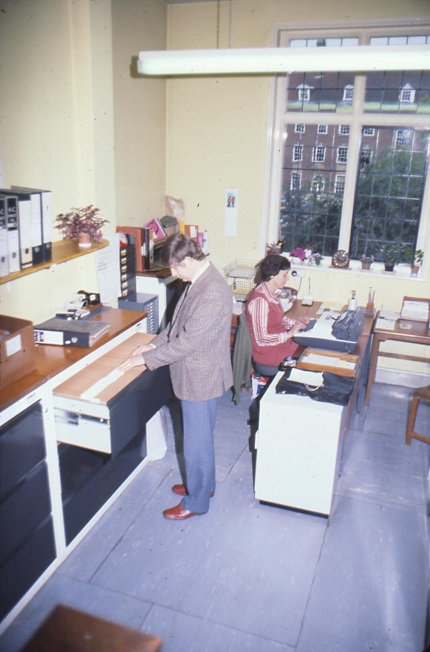 Kim Leslie is looking through a filing cabinet, Viv Hodnett is working at a typewriter, County Hall is visible through the window