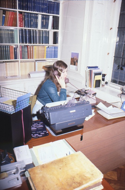 Woman talking on desk telephone, seated behind a large typewriter