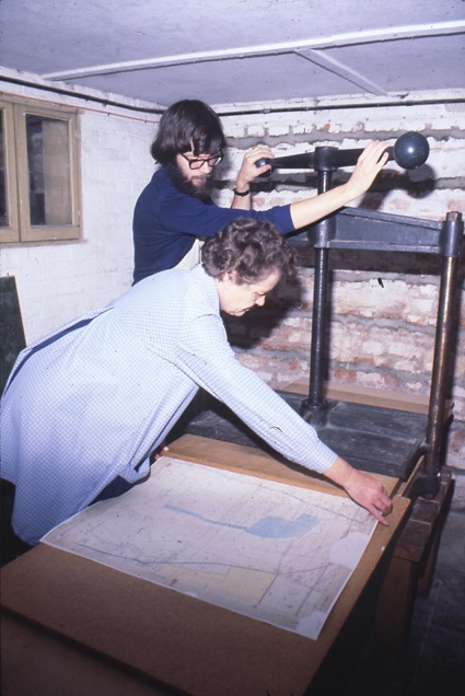 Woman leaning over and holding the edges of a map while a man winds the mechanism of a large press