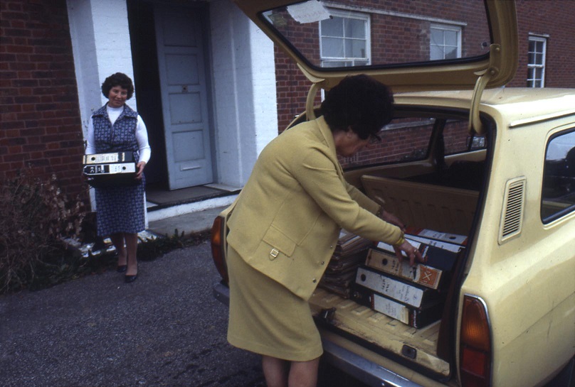 One Clerk loading files into the boot of a car, another holding files ready to be loaded into the car