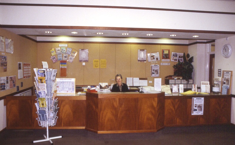 Colour photograph of Jean sitting behind a large wooden desk that spans the length of reception. Only her upper chest and upwards is visible.