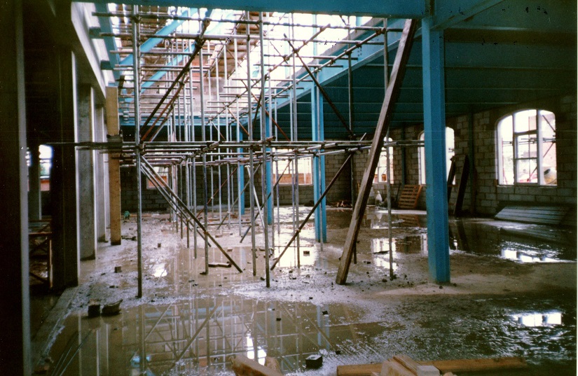Colour photograph showing the interior of the searchroom with brickwork but no roof complete, there is scaffolding up to the first floor level and many puddles on the floor