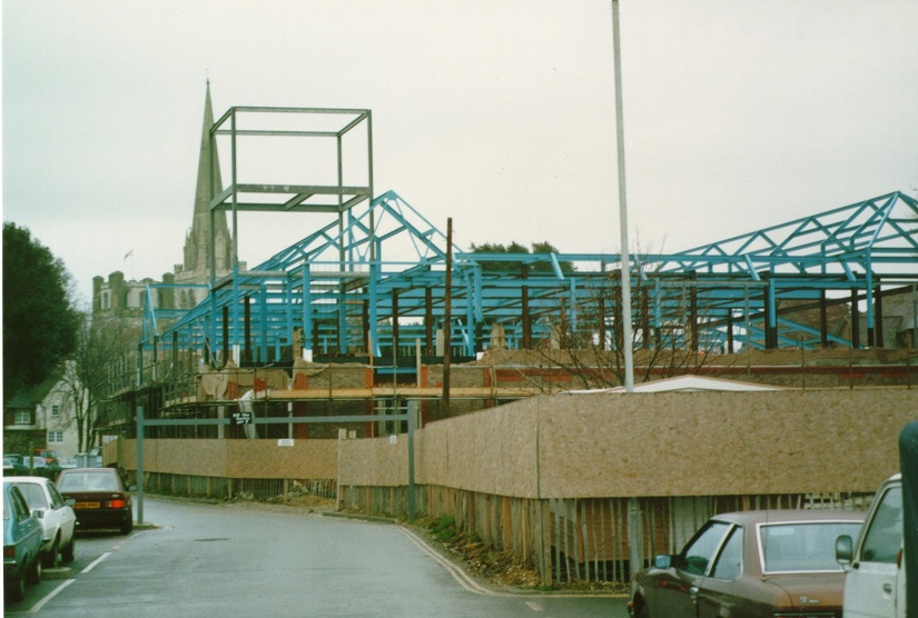 Colour photograph showing framework of building in place, shows steel frames and some brickwork, most of the ground floor is surrounding by hoarding, the Cathedral spire is visible in the background
