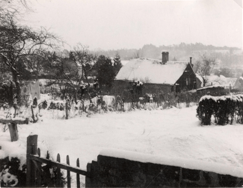 Landscape view of a snowy field. A cottage with snow on the roof, and some barren trees, as well as snow covered hillsides, are visible.