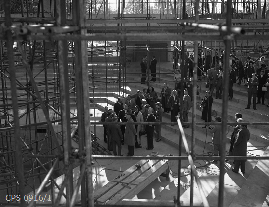 A black and white photograph taken internally from high up in the scaffolding of the roof on Chichester Festival theatre and looking down onto a crowd of visitors below.