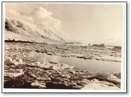 Sepia photograph of a white landscape. Icy water pools around slush and snow, and a large white hill is in the background.