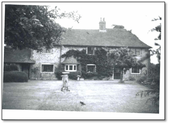 Black and white photo of the front of a cottage. Vines climb up the walls, and the front lawn is neatly maintained.