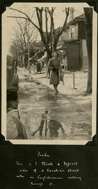A smiling woman walks down a waterlogged pavement. It is winter, and snow and ice sits on the sides next to shops and buildings.