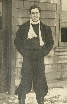 Black and white full length portrait. Tony has a white scarf on and large black trousers tucked into snow boots. He is smiling, and what appears to be a cigarette is in his mouth.