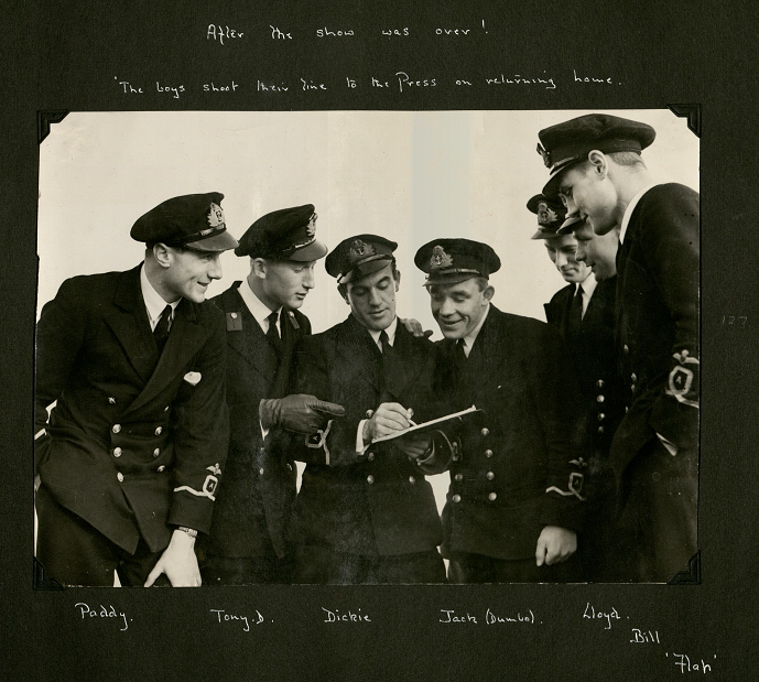 Seven men in Naval uniform crowd around and writing on a clipboard. Copyright ILN.