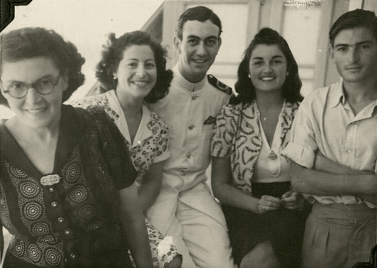 Black and white photo of five people - three women and two men. In the centre is Tony in white Naval uniform. On the far right is Don, wearing a cotton short sleeved shirt and linen trousers. The women have their hair curled and are wearing printed blouses and light cardigans.