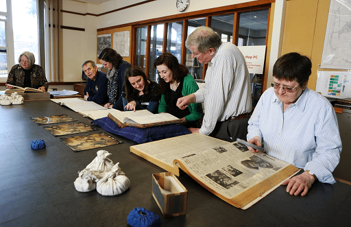 Colour photograph of seven people viewing documents on a large black table. Cloth weights are visible, as are bound newspaper volumes and aerial photographs.