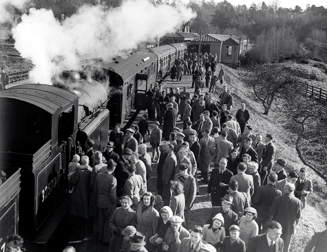 Black and white photograph. Crowds on a train station platform queue to get on and off a steam engine train.