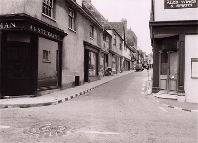 Black and white photograph of a T-junction. Two shops on the corner and a view up the narrow road.