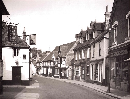 Black and white photograph showing the right hand side of the road and up featuring signs sticking out from buildings and Tudor style buildings with white paint and black beams.