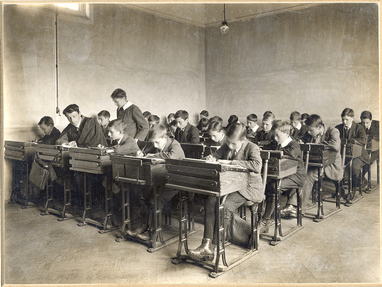 Black and white photograph of school boys working at individual desks, five rows of five in total. Some faces are blurred, as if they moved when the photo was taken. The walls are bare, and a light from the ceiling is also exposed.
