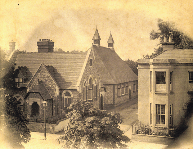 Black and white sepia toned photograph of a stone and tile roofed building on the corner of the street. The school looks a little like a chapel. Trees line the opposite side of the road.