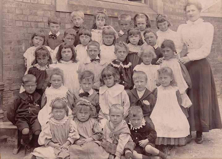 Black and white photograph of boys and girls school children, five rows of six. The school mistress stands next to the young children.