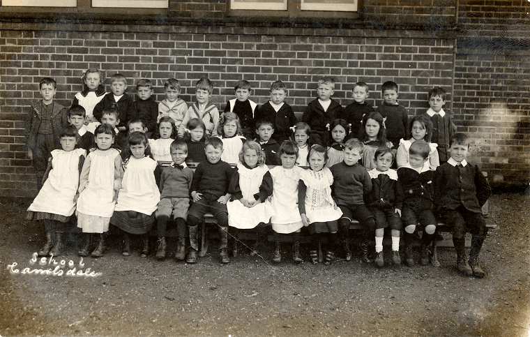 Black and white school photograph. Three rows of children, sitting, standing, and standing on benches.