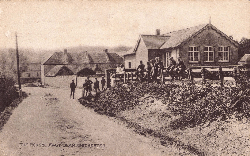 Black and white photograph. Fourteen children stand or sit on the fence outsdie the plastered school building with a tile roof, looking at the camera. There are four girls, the rest of young boys.