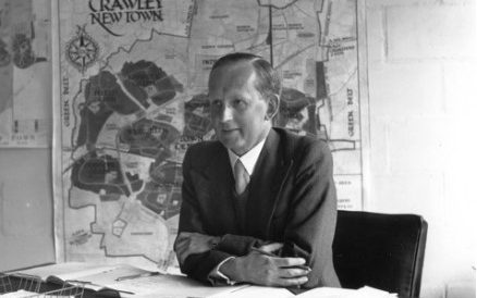 Black and white photograph of H S Howgrave Graham sat at his desk in Broadfield house, the headquarters of Crawley Development Corporation. He has the Crawley master plan pinned to the wall behind him.