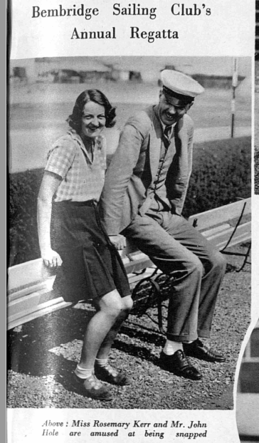 Black and white photograph of two people sitting on the back of a bench)